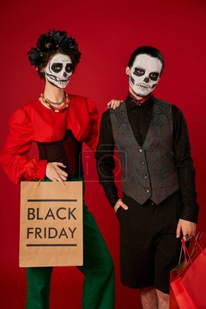 couple in dia de los muertos makeup, woman with black friday shopping bag near spooky man on red