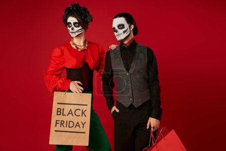 Téléchargez les photos : Couple élégant dans dia de los muertos crâne maquillage debout avec des sacs à provisions sur rouge, vendredi noir - en image libre de droit