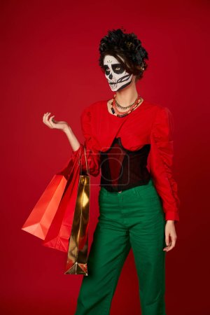 woman with spooky sugar skull makeup and shopping bags looking away on red, dia de los muertos