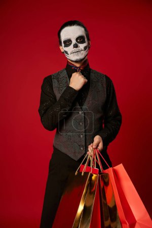 Mann in gruseligem Zuckerschädel-Make-up und festlicher Kleidung mit Einkaufstaschen auf rotem, saisonalem Verkauf