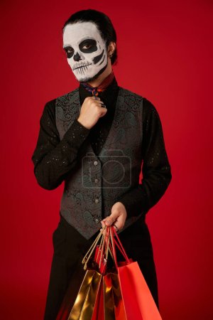 man in dia de los muertos makeup and stylish outfit posing with shopping bags on red, seasonal sale