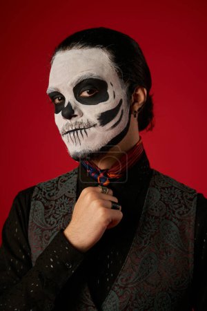 portrait of man in catrina makeup and stylish attire looking at camera on red, ria de los muertos