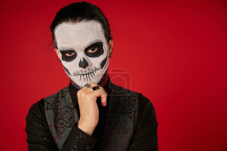 man in spooky skeleton makeup holding hand near chin and looking at camera on red, Day of Dead mug #676491212