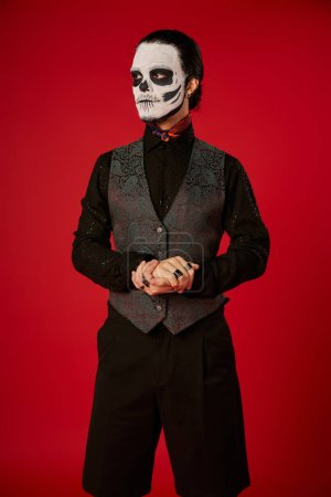 man in festive attire and sugar skull makeup looking away on red, dia de los muertos celebration