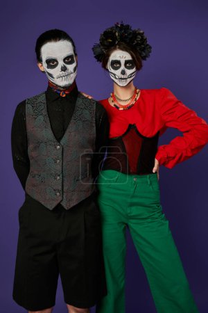 stylish couple in festive attire and dia de los muertos makeup looking at camera on blue backdrop