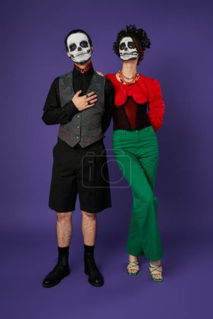 dia de los muertos couple, man in sugar skull makeup with hand on chest near spooky woman on blue