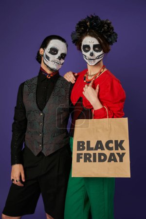 Photo for Woman in scary skull makeup and black wreath holding black friday shopping bag near man on blue - Royalty Free Image
