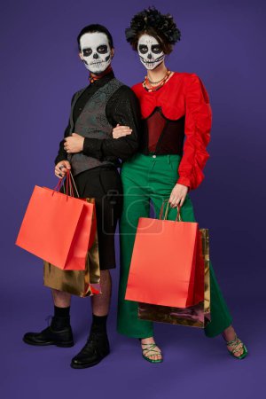 trendy dia de los muertos couple in skull makeup standing with shopping bags on blue, seasonal sale