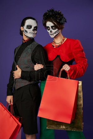 couple in dia de los muertos makeup and festive attire holding shopping bags on blue, seasonal sale