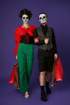 couple in dia de los muertos skull makeup and stylish attire walking with shopping bags on blue