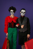 dia de los muertos couple in sugar skull makeup holding shopping bags and looking at camera on blue Poster #676491618