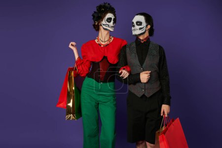Téléchargez les photos : Dia de los muertos couple en squelette maquillage tenant des sacs à provisions et se regardant sur bleu - en image libre de droit