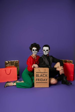 trendy couple in dia de los muertos skull makeup sitting near shopping bags on blue, black friday