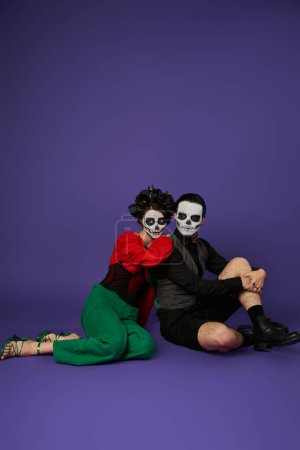 couple in traditional dia de los muertos makeup and festive attire sitting on blue, full length