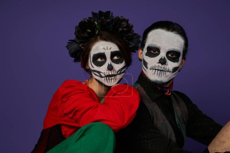 Téléchargez les photos : Dia de los muertos couple, femme dans le maquillage catrina et couronne noire appuyé sur l'homme effrayant sur bleu - en image libre de droit