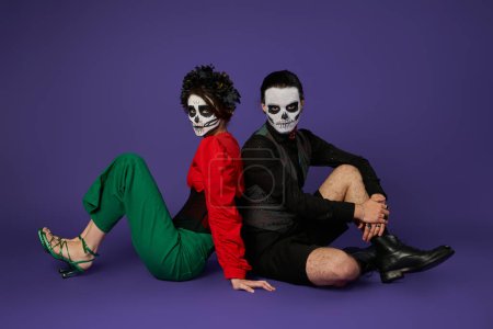 couple in sugar skull makeup and trendy outfit sitting back to back on blue, dia de los muertos fest