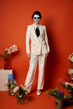 woman in skull makeup and white suit near traditional dia de los muertos ofrenda with flowers on red