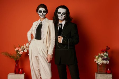 elegant couple in skull makeup and suits near altar with flowers on red, near dia de los muertos