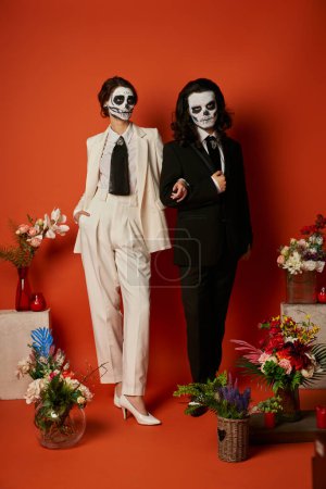 Paar in Catrina Make-up und Anzügen posiert in der Nähe von dia de los muertos ofrenda mit Blumen auf Rot