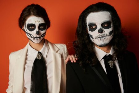 Photo for Elegant dia de los muertos couple in skull makeup, woman looking at camera near spooky man on red - Royalty Free Image