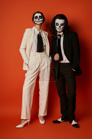 full length of stylish couple in skull makeup and elegant suits posing with hands in pockets on red