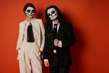 stylish couple in Day of Dead makeup, man looking at camera near woman with hands in pockets on red