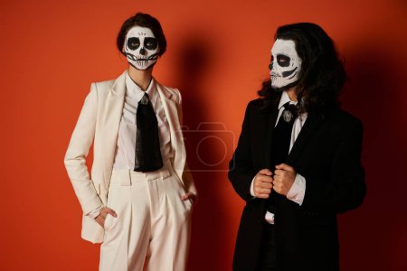 woman in sugar skull makeup and white suit standing with hands in pockets near eerie man on red