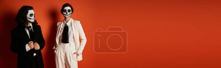 Téléchargez les photos : Femme dans le maquillage du crâne de sucre et costume blanc tenant la main dans des poches près homme effrayant sur rouge, bannière - en image libre de droit