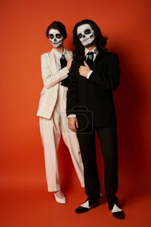 full length of couple in dia de los muertos skull makeup and festive suits looking at camera on red