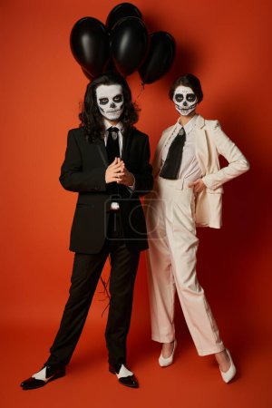 Paar in stylischen Anzügen und Catrina Calavera Make-up posiert in der Nähe schwarzer Luftballons auf Rot