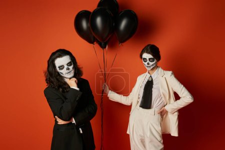Frau in Totenkopf-Make-up und weißem Anzug mit schwarzen Luftballons neben gruseligen Mann auf rot, dia de los muertos