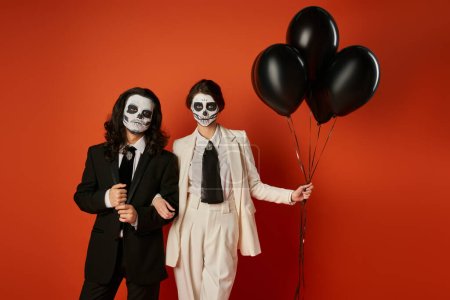 fiesta día de los muertos, elegante pareja en maquillaje de calavera de azúcar de pie con globos negros sobre rojo