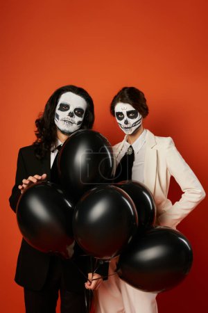 fiesta de día de los muertos, pareja espeluznante en maquillaje de cráneo mirando a la cámara cerca de globos negros sobre rojo