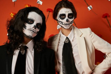 portrait of stylish couple in dia de los muertos makeup in red studio with carnation flowers decor