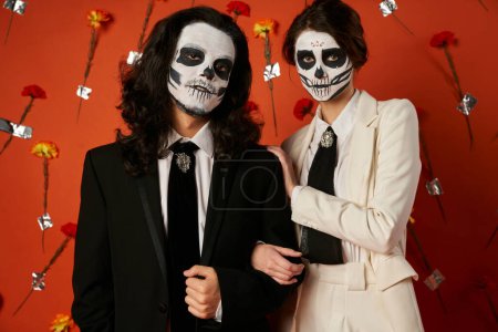 Téléchargez les photos : Couple en étrange maquillage crâne de sucre et tenue festive en regardant la caméra sur fond rouge avec des fleurs - en image libre de droit