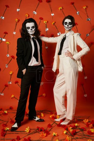 Stilvolles Paar in Totenkopf-Make-up und Anzügen auf rotem Hintergrund mit Nelken