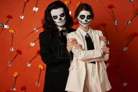 Téléchargez les photos : Dia de los muertos couple, femme avec les bras croisés près homme effrayant sur fond rouge avec des fleurs - en image libre de droit