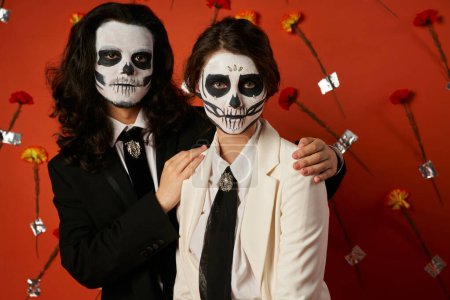 scary man hugging shoulders of woman in white suit, dia de los muertos couple on red floral backdrop