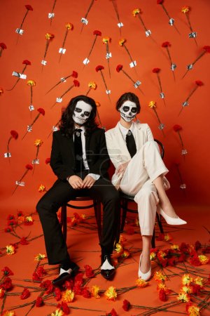Stilvolles Paar in Totenkopf-Make-up und Anzügen posiert auf Stühlen vor rotem Hintergrund mit Blumen, Day of Dead