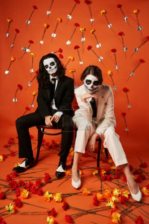 trendy couple in skull makeup and suits looking at camera on chairs on red backdrop with carnations