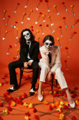 trendy couple in skull makeup and suits looking at camera on chairs on red backdrop with carnations puzzle #676495214