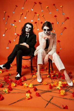 full length of elegant couple in skull makeup and suits sitting on chairs in red studio with flowers