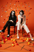 full length of elegant couple in skull makeup and suits sitting on chairs in red studio with flowers hoodie #676495234