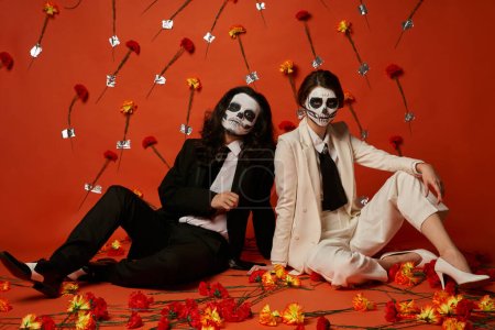Paar in Zuckerschädel-Make-up und eleganten Anzügen sitzt auf dem Boden in rotem Studio mit Nelkenblüten