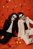 couple in scary catrina makeup and suits sitting on floor in red studio with carnation flowers puzzle #676495336