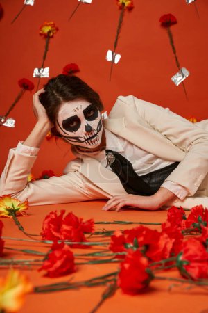 Foto de Mujer elegante en maquillaje de cráneo de azúcar y traje blanco acostado cerca de claveles en estudio rojo - Imagen libre de derechos