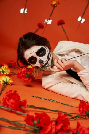 woman in catrina makeup and suit lying down and looking at camera near carnations on red backdrop