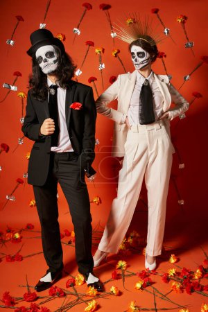 Foto de Fiesta día de los muertos, pareja en esqueleto maquillaje y atuendo festivo en estudio rojo con claveles - Imagen libre de derechos