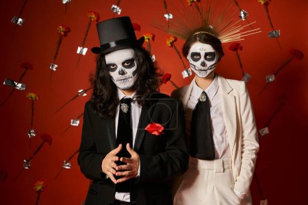 dia de los muertos party, couple in  scary makeup looking at camera in red studio with carnations