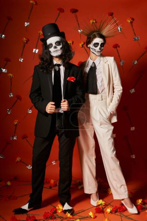 full length of elegant couple in dia de los muertos makeup on red backdrop with floral decor